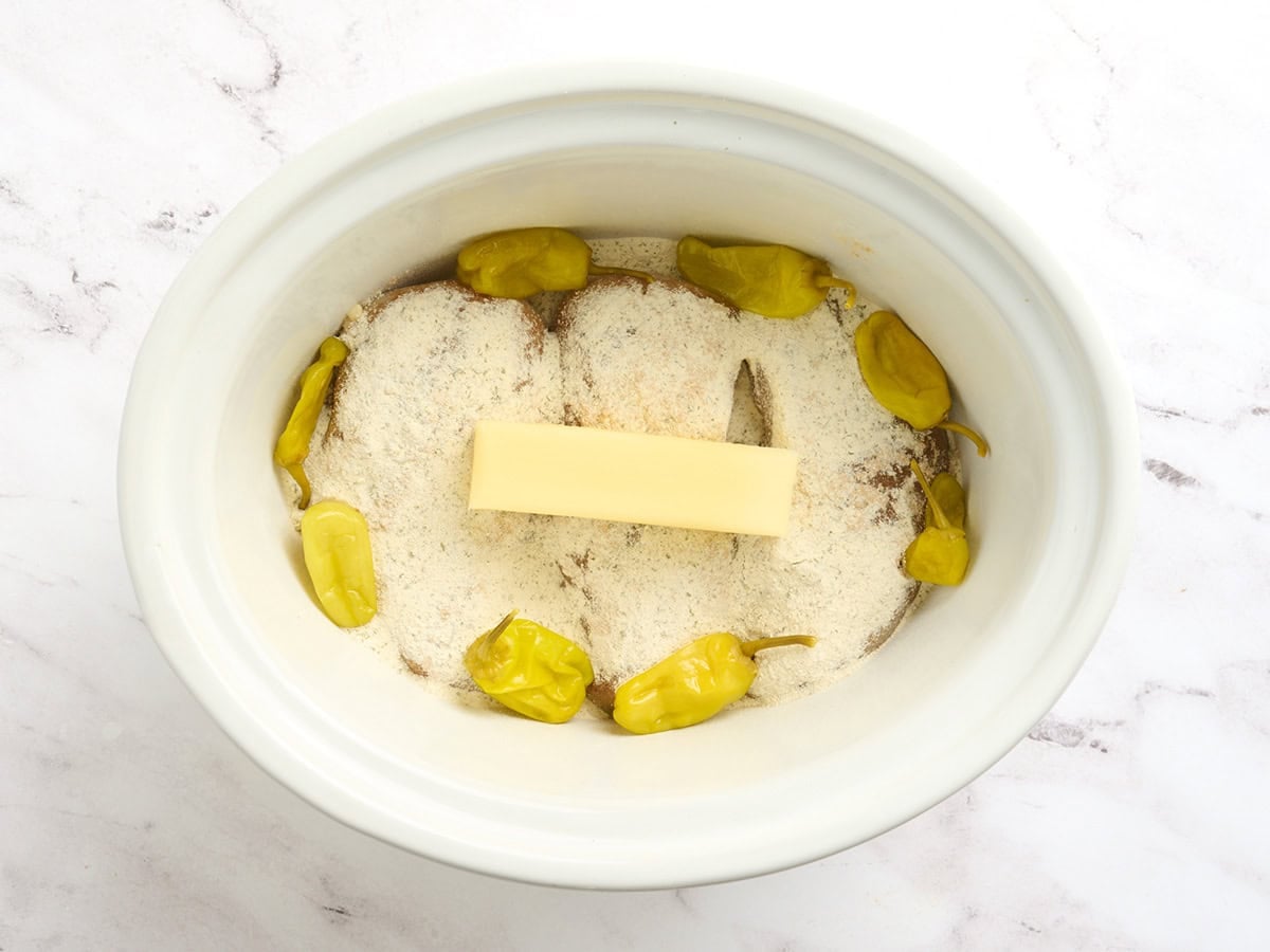Chicken breasts in a crockpot with seasonings, a stick of butter, and pepperoncini peppers to make Mississippi chicken.