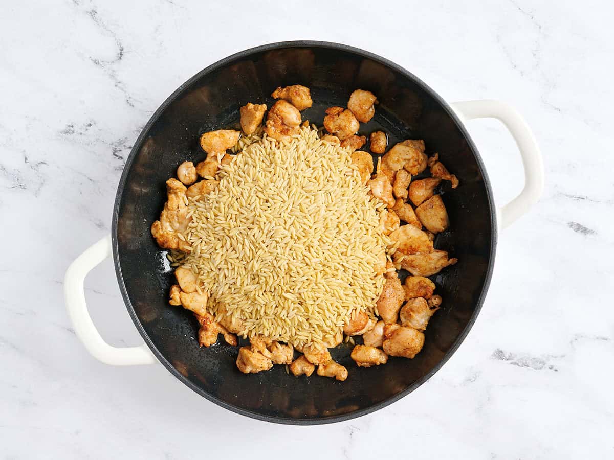 Uncooked orzo added to a skillet with cooked chicken.