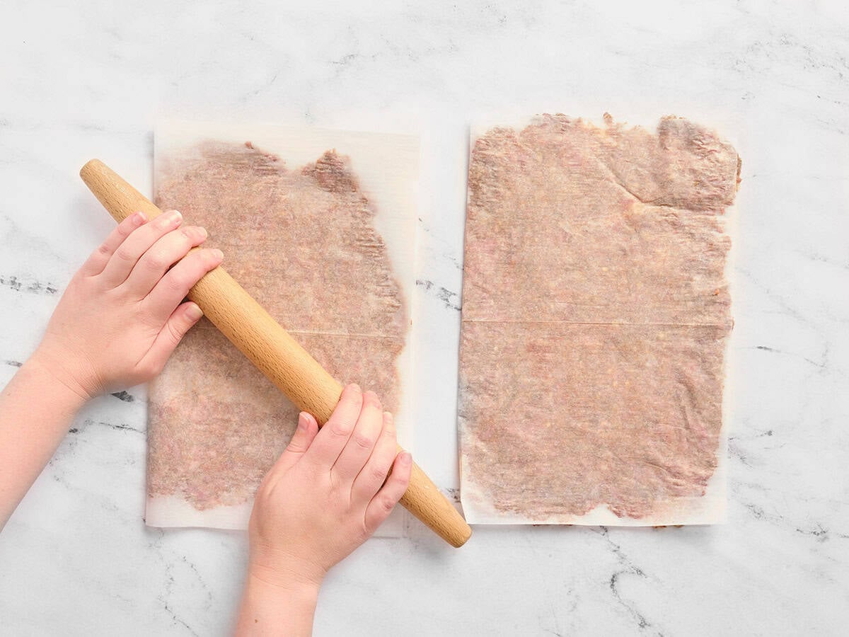 Hands using a rolling pin to flatten ground beef between two pieces of parchment paper until flattened.