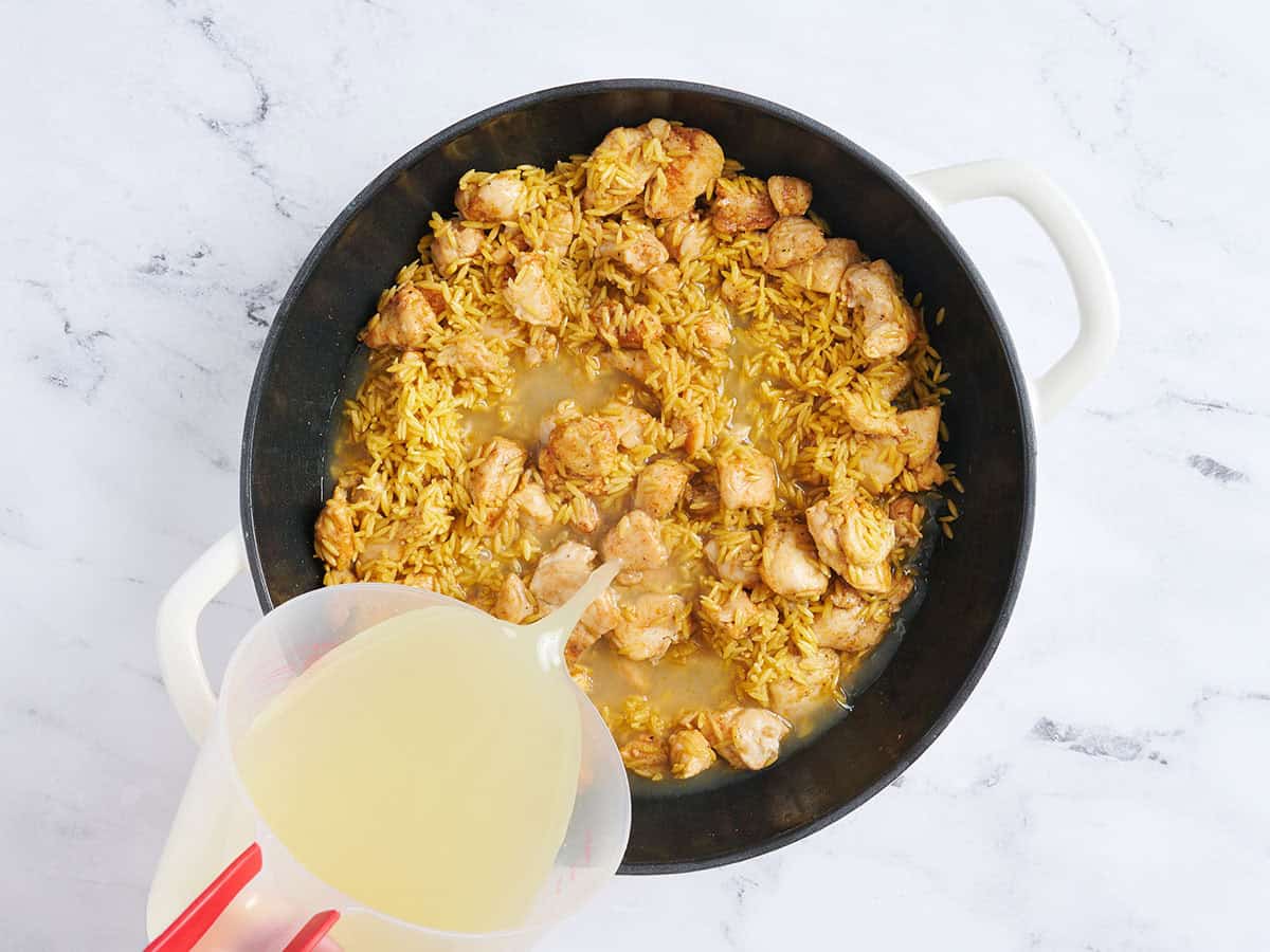 Broth being poured into a skillet with cooked chicken and toasted orzo.