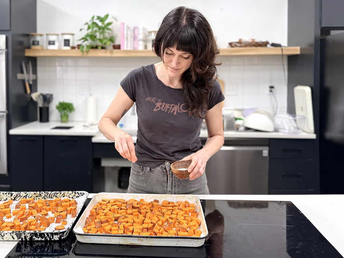 A women sprinkling salt over a sheet pan of diced sweet potatoes in a kitchen.