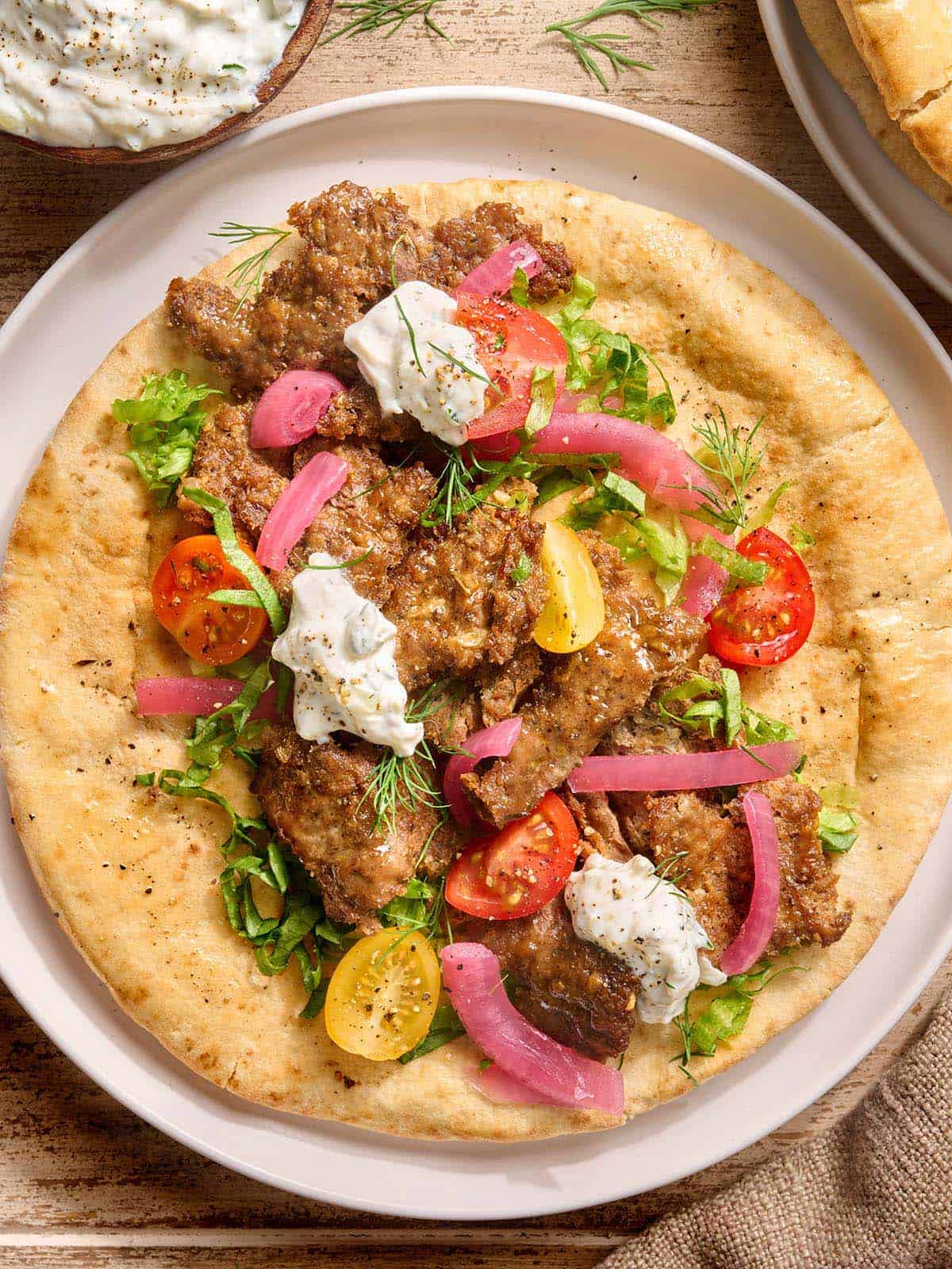 Overhead view of a sheet pan doner kebab on a pita bead with salad and tzatziki sauce.