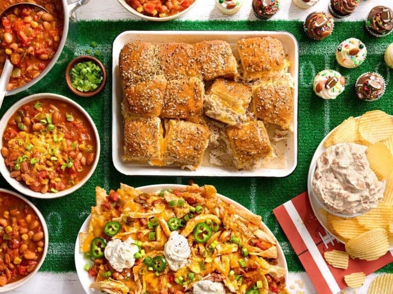 Overhead view of a game day spread, including chicken sliders, chili, nachos, dip and cup cakes.