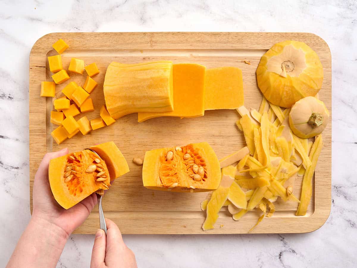 Hands prepping butternut squash by removing the seeds, surrounded by butternut squash peel and diced butternut squash on a cutting board.