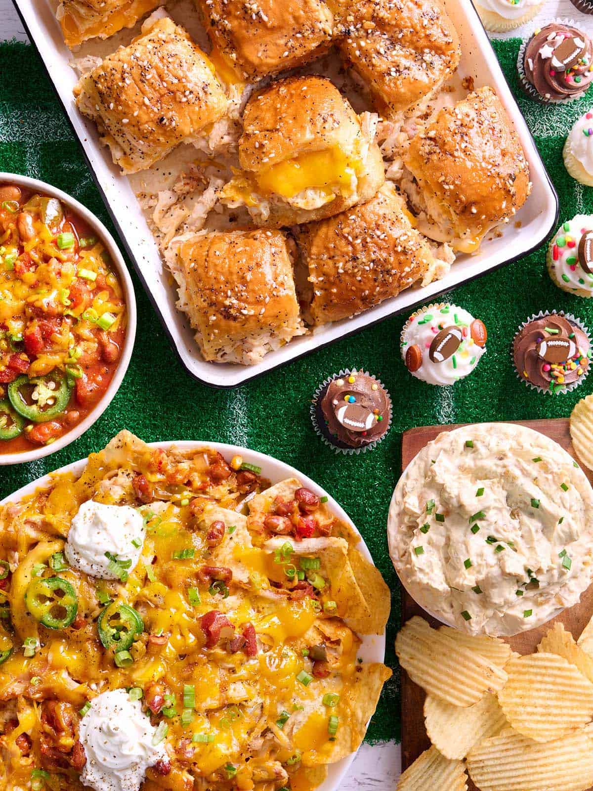 Overhead view of a game day spread, including chicken sliders, chili, nachos, dip and cup cakes.