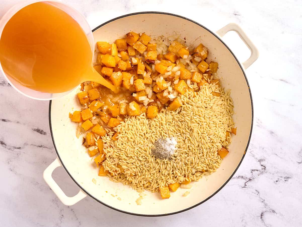 Butternute squash in a skillet with orzo and seasonigs, with broth being poured in.
