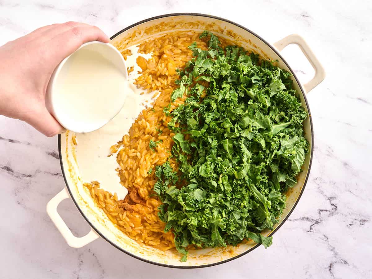 Butternut squash orzo in a skillet with fresh kale and cream being poured in.