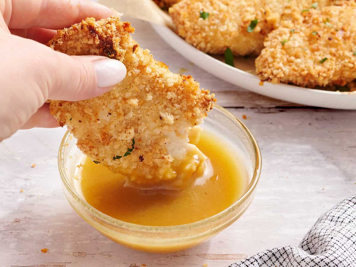 A hand dipping half a chicken tender in honey mustard sauce.