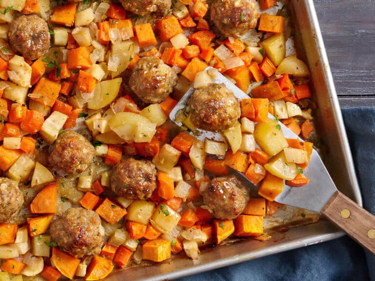 Overhead view of sheet pan turkey meatballs and root vegetables with a metal spatula.