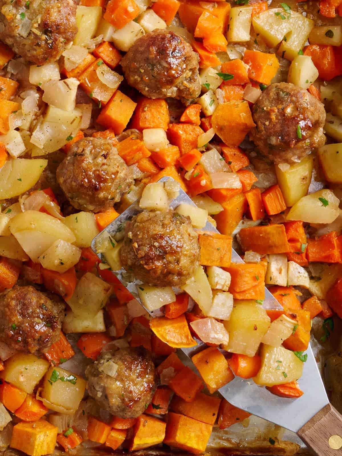 Overhead view of sheet pan turkey meatballs and root vegetables with a metal spatula.
